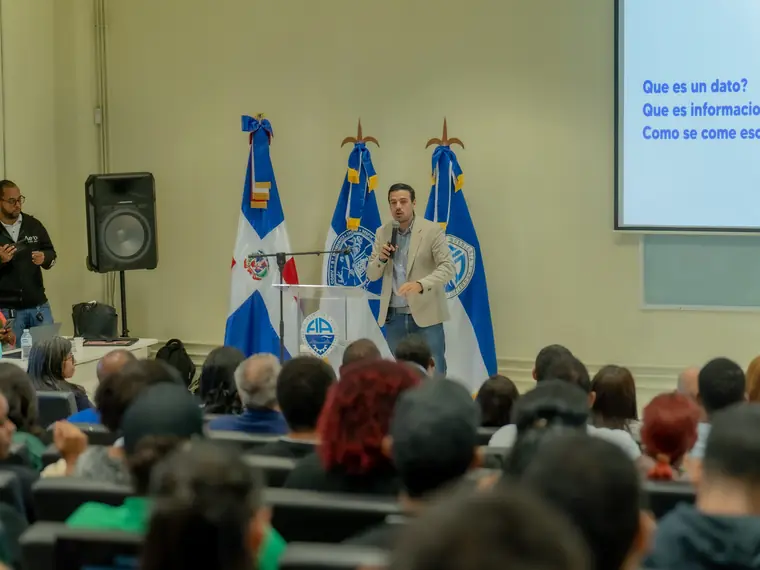 Auditorio universitario lleno de público durante una charla, con banderas dominicanas al fondo del escenario