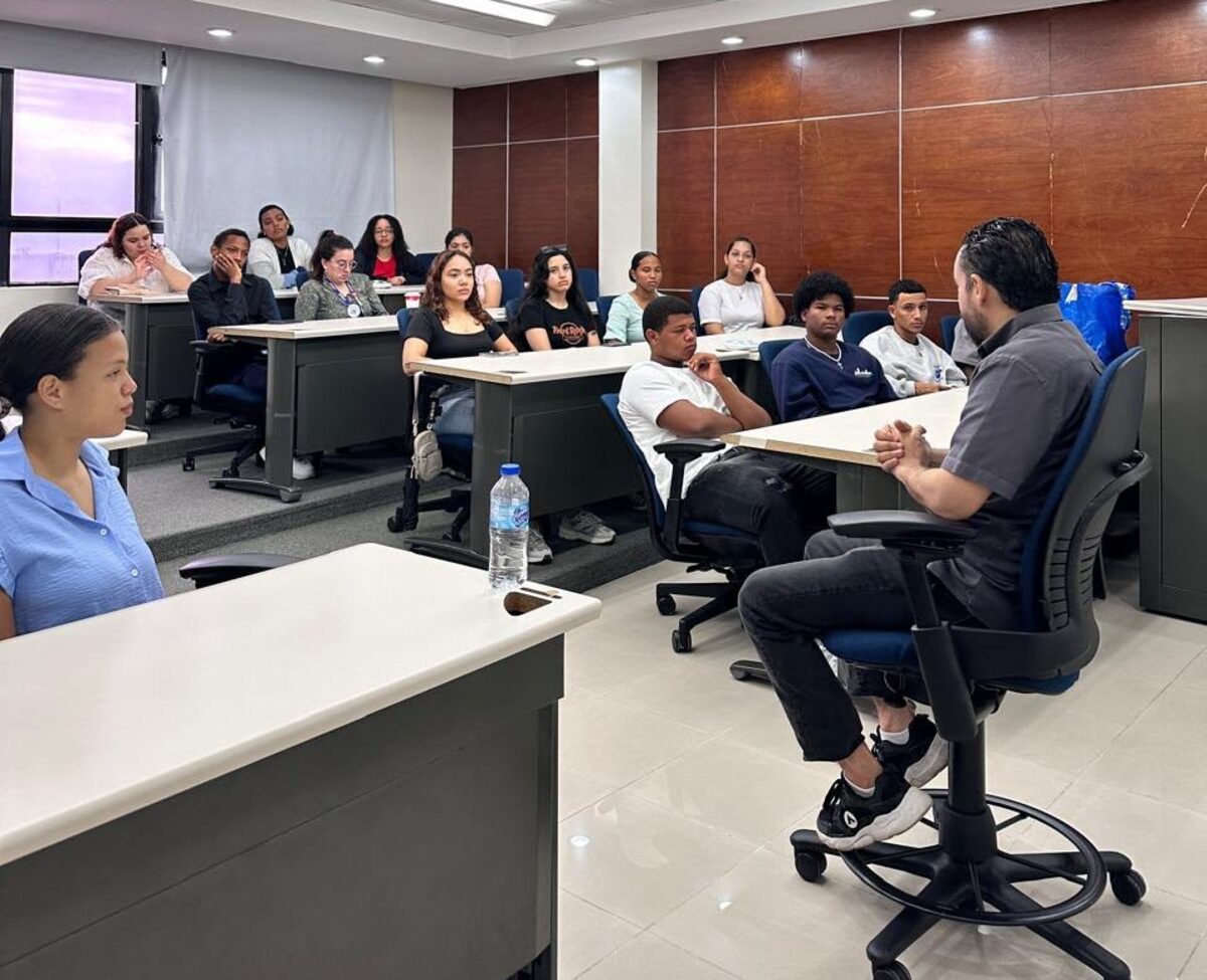 Estudiantes de secundaria sentados en pupitres escuchan a un facilitador que conversa con ellos en el aula.