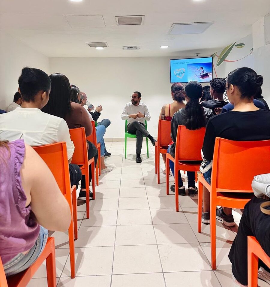 Un facilitador conversa sentado frente a un grupo de jóvenes en un salón, con una presentación proyectada al fondo.